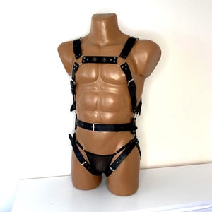 May include: Black leather body harness with adjustable straps and metal hardware. The harness includes a chest piece, shoulder straps, and a waist belt. A matching black mesh brief is also included. The harness is displayed on a mannequin.