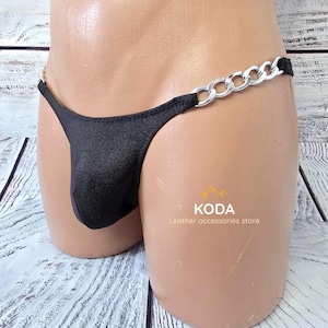 May include: A black thong featuring a metallic silver chain on the side. The garment is displayed on a mannequin torso. The text KODA Leather accessories store is visible on the lower right side of the black fabric portion of the garment.