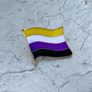 May include: A gold-colored pin with a flag design. The flag has five horizontal stripes: yellow, white, purple, white, and black. The flag is slightly wavy.