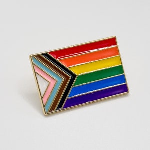 May include: A gold-toned enamel pin featuring a rainbow flag with a black, brown, light blue, pink, and white chevron design.