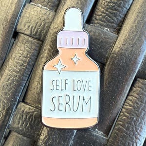 May include: A white enamel pin shaped like a bottle of serum with a pink lid and a peach colored body. The bottle has two white stars and the text "SELF LOVE SERUM" in black.