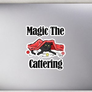 May include: A sticker with a black cat under a red blanket with the text "Magic The Cattening" on a silver laptop.