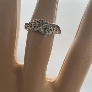 May include: A silver ring featuring a leaf design. The ring has two detailed leaves, with textured veins, set side by side. The ring is shown on a finger against a white background.
