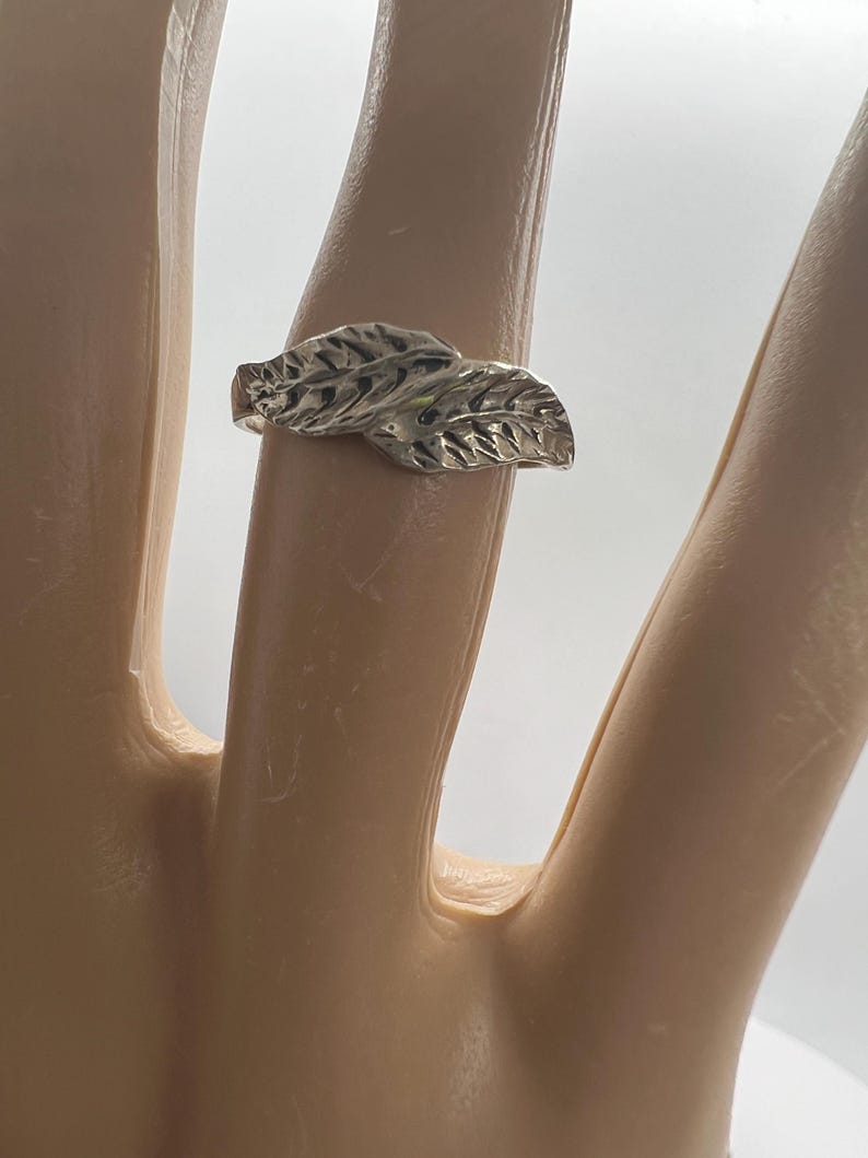 May include: A silver ring featuring a leaf design. The ring has two detailed leaves, with textured veins, set side by side. The ring is shown on a finger against a white background.