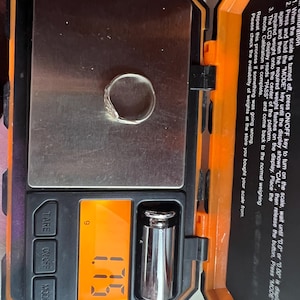 May include: A silver ring with a decorative design is placed on a digital scale. The scale displays a weight of 1.7 grams. The scale is black and orange, with buttons for TARE, ON/OFF, and MODE.