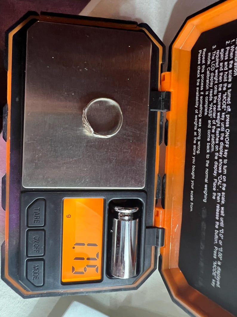 May include: A silver ring with a decorative design is placed on a digital scale. The scale displays a weight of 1.7 grams. The scale is black and orange, with buttons for TARE, ON/OFF, and MODE.