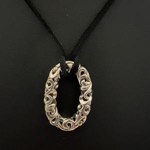 May include: Silver-toned oval pendant with an intricate, swirling design, suspended from a black cord. The pendant's openwork pattern creates a delicate, ornate appearance. The background is a solid black.