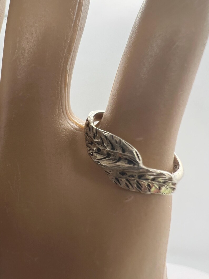 May include: A silver ring featuring a leaf design. The ring is worn on a finger, showcasing the intricate details of the leaf's texture. The ring's band is smooth and polished, contrasting with the textured leaf.