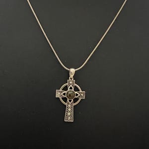 May include: Silver-toned Celtic cross pendant necklace. The cross features a central dark stone and is adorned with small, decorative accents. The necklace hangs on a delicate silver chain, set against a dark background.