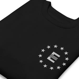 Puede incluir: Sudadera negra con cuello redondo. Presenta un diseño bordado en blanco de un cuadrado con líneas horizontales en el interior, rodeado de estrellas blancas. El diseño está en el pecho izquierdo.