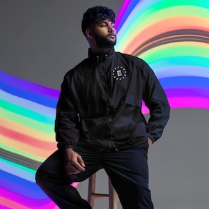 May include: A black windbreaker jacket with a zip fastening and a white embroidered logo featuring the letter "E" surrounded by stars. The jacket is being worn by a person with a beard and a black shirt. The background is a blurred rainbow.