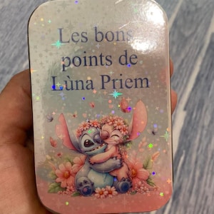 Peut inclure: Une boîte rectangulaire argentée avec une illustration colorée de deux personnages de dessins animés s'embrassant au milieu de fleurs. La boîte porte l'inscription "Les bons points de Luna Priem" en bleu. Le design comprend des papillons et des paillettes.