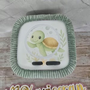 Peut inclure: Un objet carré blanc avec une bordure en peluche verte. Le centre présente une illustration à l'aquarelle d'une tortue verte souriante avec une carapace marron. Le design comprend des motifs floraux et deux boutons noirs ovales.