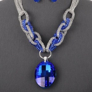 May include: A silver chain necklace with a large blue crystal pendant and matching earrings. The necklace is made of multiple strands of chain and features a large, oval-shaped pendant with a faceted blue crystal.