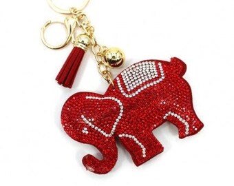 May include: A red elephant keychain with a gold-colored chain, ring, and tassel. The elephant is covered in red rhinestones with white rhinestone accents. A small gold bell hangs from the chain.
