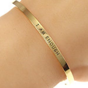 May include: A gold-colored metal cuff bracelet. The bracelet is engraved with the words "I AM ENOUGH" in a simple, sans-serif font. The bracelet is shown on a person's wrist.