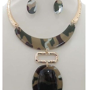 May include: A gold and camouflage patterned necklace with a large oval pendant. The pendant has a black center and a gold frame. The necklace is made of plastic and metal.