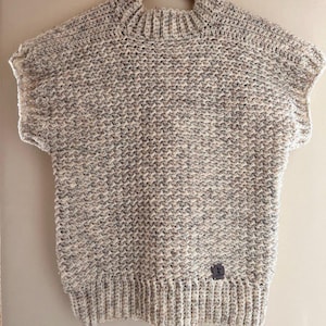 Puede incluir: Un suéter beige sin mangas de ganchillo con cuello alto y una pequeña etiqueta marrón en la parte inferior derecha.