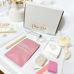 Könnte beinhalten: Eine Sammlung von Gegenständen, darunter eine weiße Schachtel mit Text, eine Spritze, ein rosa Päckchen mit deutschem Text, ein kleiner klarer Behälter, ein Holzstab und andere kleine Gegenstände. Die Gegenstände sind auf einer weißen Oberfläche angeordnet.