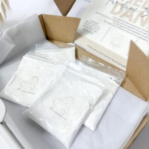 Peut inclure: Une boîte en carton ouverte contenant plusieurs petits sacs en plastique transparents remplis de poudre blanche. Les sacs sont scellés et portent un logo imprimé. La boîte contient également des cartes imprimées et du papier de soie.