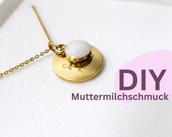 May include: A gold-toned necklace featuring a circular pendant with a smooth, white, opaque cabochon centre. The pendant is engraved with small, thin symbols. The image includes the text DIY Muttermilchschmuck in purple on a light pink background.