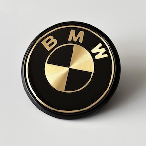 May include: A circular emblem featuring the BMW logo in a black and gold color scheme. The design includes the letters BMW at the top, with a central quadrant pattern alternating between brushed gold and black sections, all enclosed by a gold border.