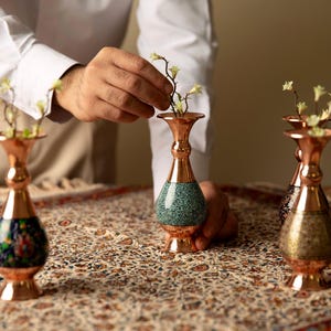 May include: Copper vases with floral designs and turquoise accents. The vases are arranged on a patterned tablecloth, with a person's hands arranging flowers in one of the vases. The vases are of varying heights and designs.