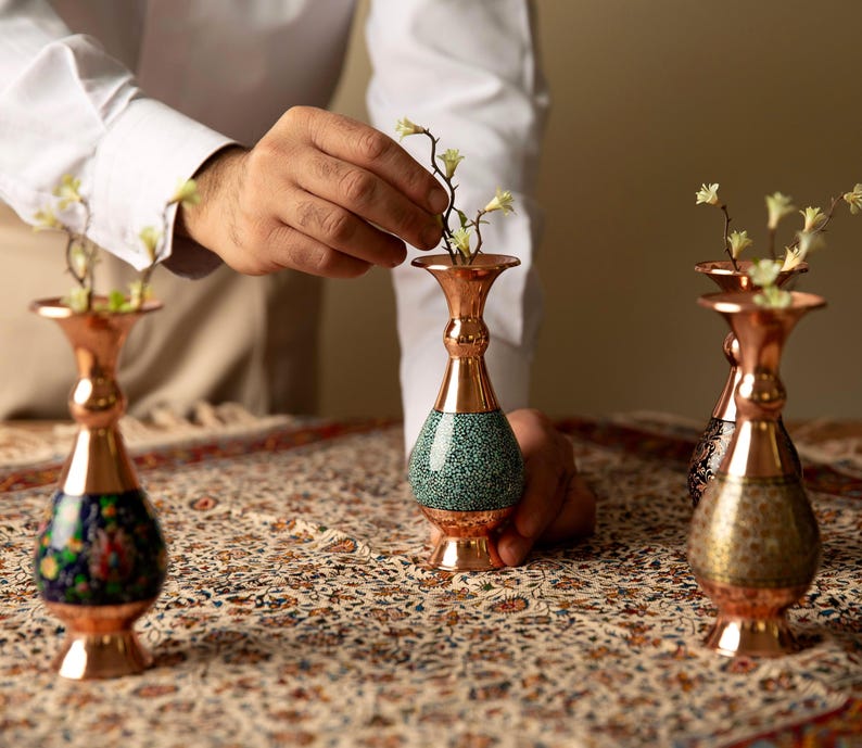 May include: Copper vases with floral designs and turquoise accents. The vases are arranged on a patterned tablecloth, with a person's hands arranging flowers in one of the vases. The vases are of varying heights and designs.