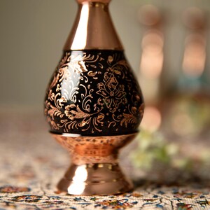 May include: A copper-colored vase with a black, floral-patterned center. The vase has a bulbous shape with a flared base and a narrow neck. The floral design is in a lighter copper color, creating a contrast against the black background.