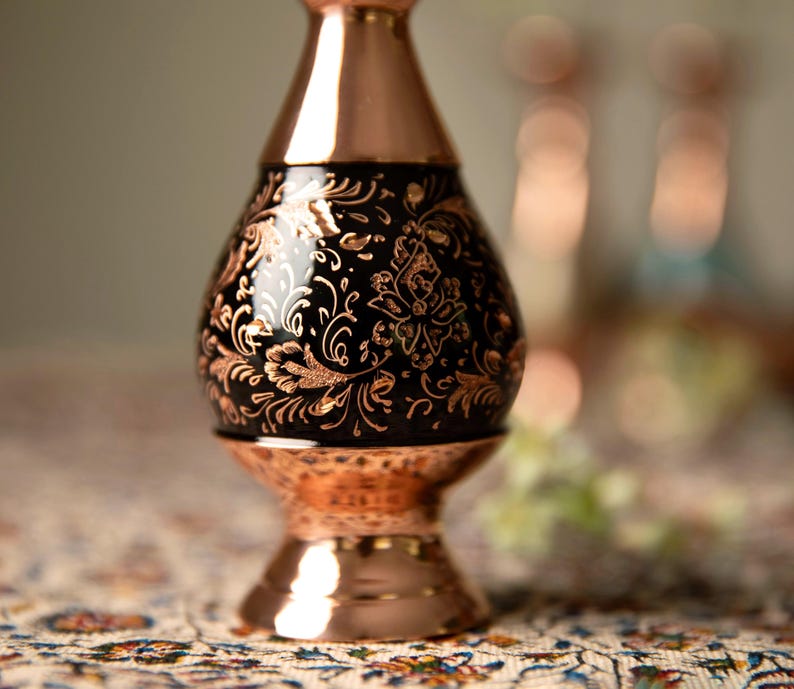 May include: A copper-colored vase with a black, floral-patterned center. The vase has a bulbous shape with a flared base and a narrow neck. The floral design is in a lighter copper color, creating a contrast against the black background.