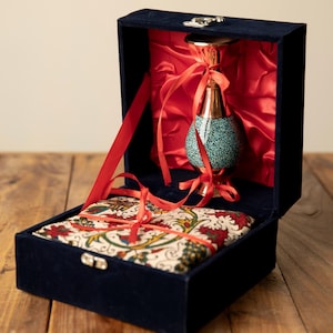 May include: A navy blue velvet box with a decorative vase and a patterned fabric. The vase has a copper-colored top and base with a turquoise speckled body. Red ribbons and lining add contrast. The box is open, revealing the contents.