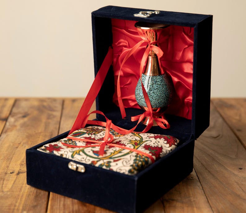 May include: A navy blue velvet box with a decorative vase and a patterned fabric. The vase has a copper-colored top and base with a turquoise speckled body. Red ribbons and lining add contrast. The box is open, revealing the contents.