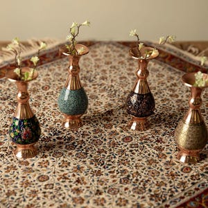 May include: Four copper vases with floral designs and a flared top, each holding small sprigs of white flowers. The vases have a bulbous base with different patterns: blue floral, green speckled, black floral, and gold patterned.