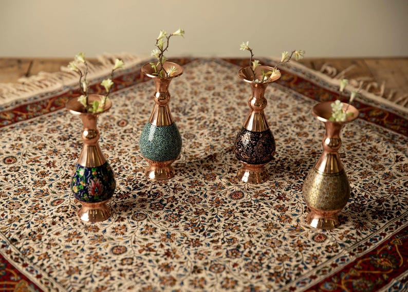 May include: Four copper vases with floral designs and a flared top, each holding small sprigs of white flowers. The vases have a bulbous base with different patterns: blue floral, green speckled, black floral, and gold patterned.