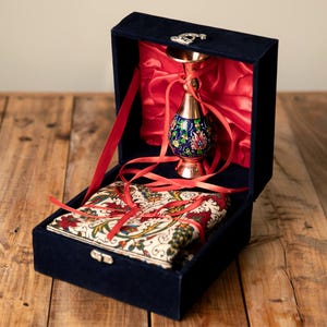 May include: A navy blue velvet box with a decorative metal clasp, containing a copper vase with a blue floral design and a patterned fabric. The box is lined with red satin and tied with red ribbons.