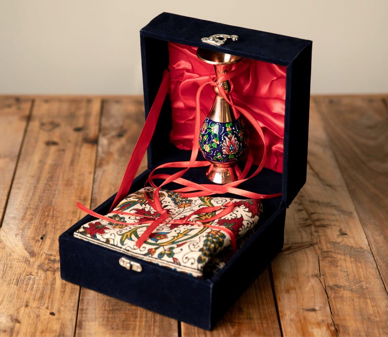 May include: A navy blue velvet box with a decorative metal clasp, containing a copper vase with a blue floral design and a patterned fabric. The box is lined with red satin and tied with red ribbons.