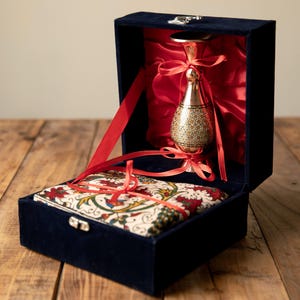 May include: A navy blue velvet box with a red satin interior holds a decorative gold-colored metal vessel and a patterned fabric item, both tied with red ribbons. The box is open, revealing the contents.