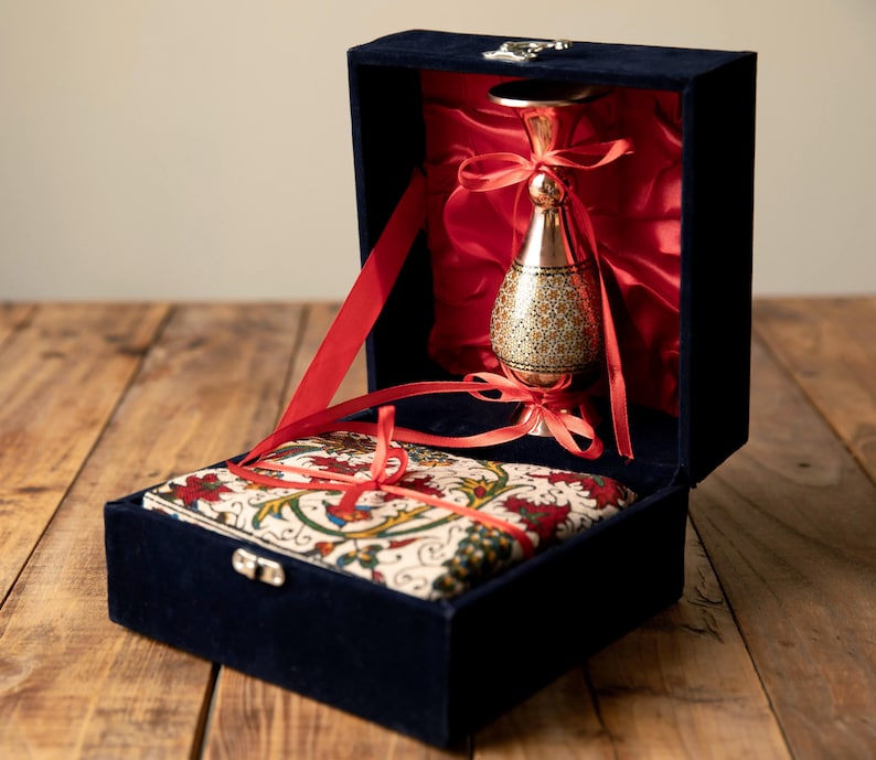 May include: A navy blue velvet box with a red satin interior holds a decorative gold-colored metal vessel and a patterned fabric item, both tied with red ribbons. The box is open, revealing the contents.