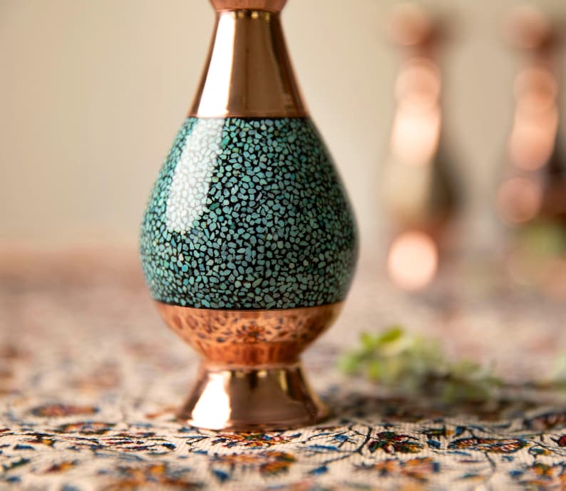 May include: A copper vase with a turquoise mosaic design on the body. The vase has a flared base and a narrow neck, with a polished copper finish. The vase is likely handmade and is a decorative item.