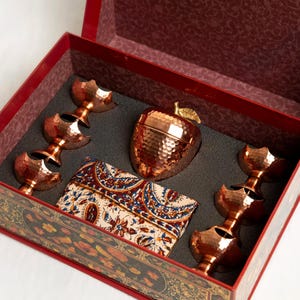 Può includere: Un set regalo in una scatola rossa contenente un contenitore in rame martellato a forma di mela con stelo dorato, sei piccole tazze in rame martellato disposte in due colonne e una pochette rettangolare in tessuto con motivo paisley blu, rosso e oro.