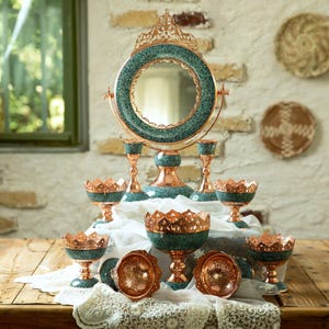 Può includere: Una collezione di oggetti decorativi in rame e turchese, tra cui uno specchio, candelabri e ciotole, disposti su una superficie di legno. Lo specchio ha una cornice rotonda con dettagli turchesi. Le ciotole e i candelabri hanno un design simile.
