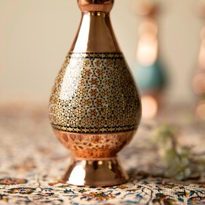 May include: A copper vase with a patterned design. The vase has a bulbous body with a detailed mosaic pattern in gold and black. The vase has a flared base and a narrow neck. The background is blurred, with other vases visible.