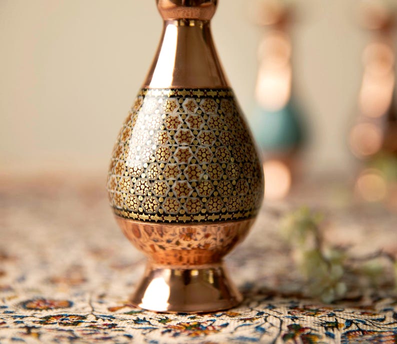 May include: A copper vase with a patterned design. The vase has a bulbous body with a detailed mosaic pattern in gold and black. The vase has a flared base and a narrow neck. The background is blurred, with other vases visible.