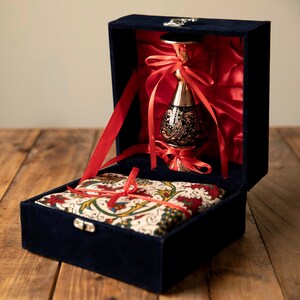 May include: A navy blue velvet box with a silver clasp, opened to reveal a decorative black and gold vase tied with red ribbons. The box also contains a patterned fabric cushion.