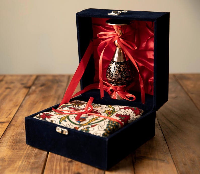 May include: A navy blue velvet box with a silver clasp, opened to reveal a decorative black and gold vase tied with red ribbons. The box also contains a patterned fabric cushion.