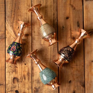 May include: Four copper vases with ornate designs. Each vase has a flared top and a bulbous, decorated midsection. The midsections feature patterns in gold, black, blue, and green. The vases are arranged on a wooden surface.
