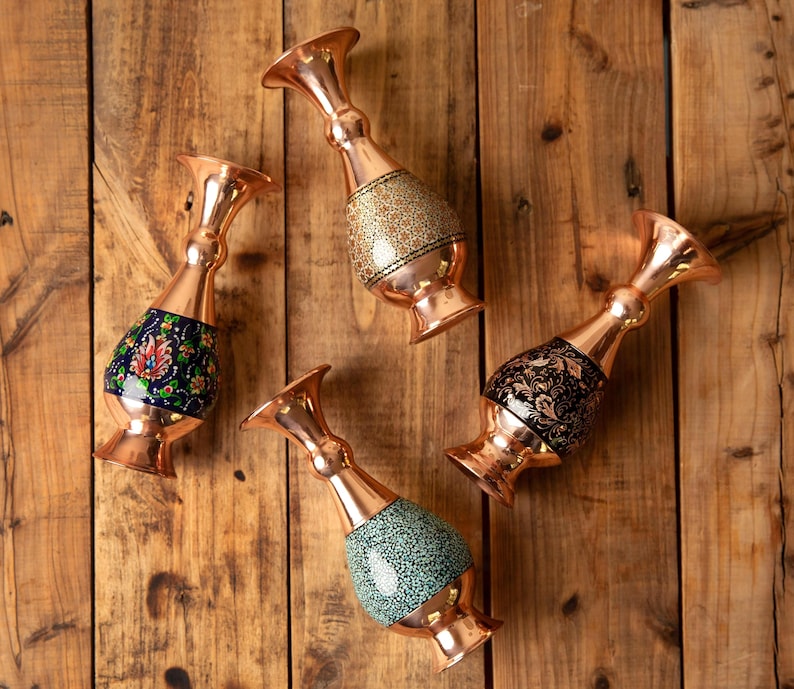 May include: Four copper vases with ornate designs. Each vase has a flared top and a bulbous, decorated midsection. The midsections feature patterns in gold, black, blue, and green. The vases are arranged on a wooden surface.