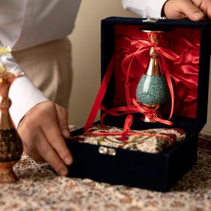 May include: A decorative vase with a turquoise speckled body and gold accents is presented in a navy blue velvet box lined with red satin. The vase is tied with red ribbons. A second copper vase is visible.