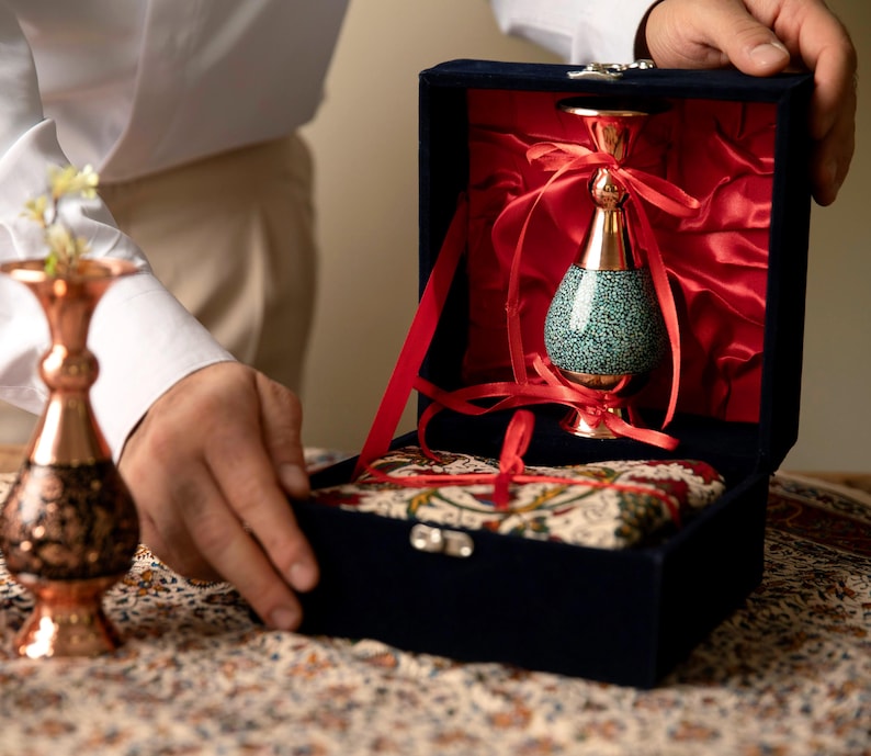 May include: A decorative vase with a turquoise speckled body and gold accents is presented in a navy blue velvet box lined with red satin. The vase is tied with red ribbons. A second copper vase is visible.