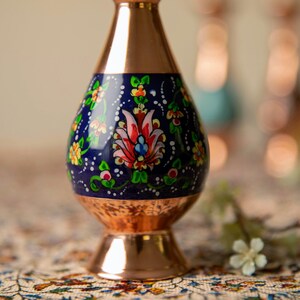 May include: A copper vase with a navy blue center section adorned with a floral design in red, pink, yellow, and green. The vase has a flared base and a narrow neck, showcasing a blend of metallic and painted elements.
